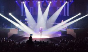 A person performs on a brightly lit stage in front of an audience, with musicians and equipment in the background.