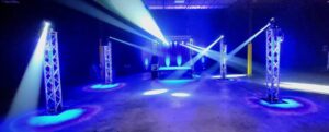 Empty event space with stage, metal trusses, and blue spotlights illuminating the area in a dark warehouse.