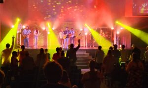 A band performs on a brightly lit stage while an audience stands and raises hands in front of them.