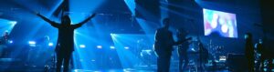 Silhouetted band performing on stage with blue lighting and a large screen displaying colorful circles in the background.