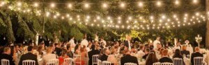 People dressed formally sit at tables outdoors under string lights for an evening event, surrounded by trees.