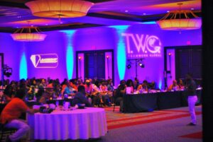 A speaker addresses a seated audience in a large conference room with “Teamwork Global” projected on the wall.
