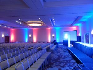 Empty conference room with rows of chairs, a lit stage, and red and blue uplighting along the walls.