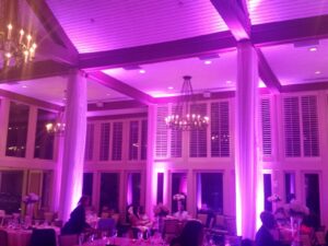 A banquet hall with purple lighting, set dining tables, chandeliers, and tall windows with shutters at night.