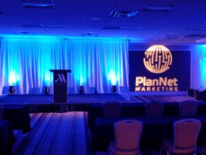 Stage with podium and PlanNet Marketing logo projection, blue lighting, and rows of empty chairs in conference room.