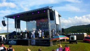 Outdoor music festival stage with band performing; audience seated on grass, umbrellas, and portable toilets visible.