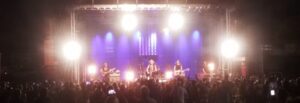 A band performs on a brightly lit outdoor stage at night, with a crowd of people watching in the foreground.