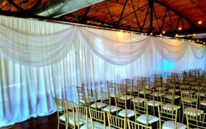 Rows of gold chairs are arranged in front of white draped curtains under exposed wooden beams and string lights.