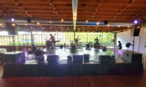 A band is performing on an indoor stage with instruments and speakers, under exposed wooden beams and string lights.