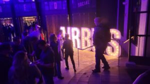 People gather near a large, illuminated “FORBES” sign at an indoor event with purple lighting.