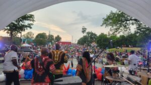 Musicians perform on stage at an outdoor event, facing a large seated audience under a cloudy sky.