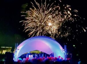 Fireworks light up the night sky above a white domed stage decorated with balloons and illuminated by colored lights.