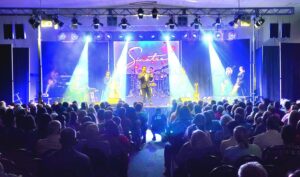 A band performs on stage with bright lights and a crowd seated in front; “Sinatra” is displayed behind the performers.