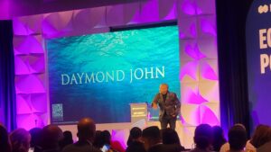 Daymond John stands on stage speaking at an event, with his name displayed on a large screen behind him.