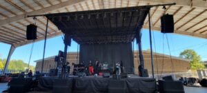 A band performs soundcheck on an outdoor covered stage with equipment and speakers visible around them.