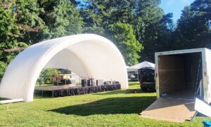 White inflatable stage tent and sound equipment setup on grass, with a trailer and trees in the background.