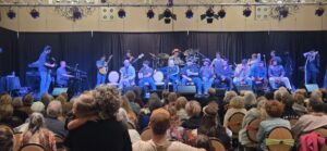 A group of people sits on stage facing an audience, with musical instruments and equipment arranged behind them.