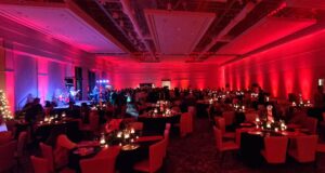 Large banquet hall with red lighting, decorated tables, and people gathered near a stage with a live band performing.