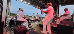 A band dressed in white performs on an outdoor stage in front of a large crowd during daylight.