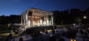 Outdoor concert at dusk with musicians performing on stage and audience seated at round tables under evening sky.