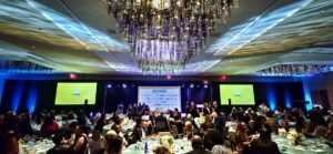 Large banquet hall filled with seated guests, decorated tables, and a stage with screens and banners at the front.