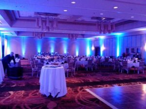 Large banquet hall with round tables set for an event, blue uplighting, and one person arranging items.