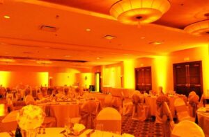 Large banquet hall with round tables, covered chairs, floral centerpieces, and warm yellow-orange lighting.