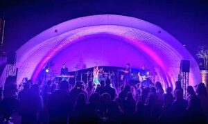 A band performs on a lit stage with a curved canopy at night as a crowd watches.