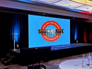 Stage with large screen displays “Shock & Awe Productions” logo; round tables and water bottles in foreground.