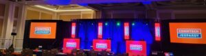 Large conference room with a stage set for “CommTech Jeopardy,” featuring screens and podiums under colorful lights.