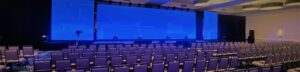 Rows of empty chairs face a large blue-lit stage in a conference room with black curtains and ceiling lights.